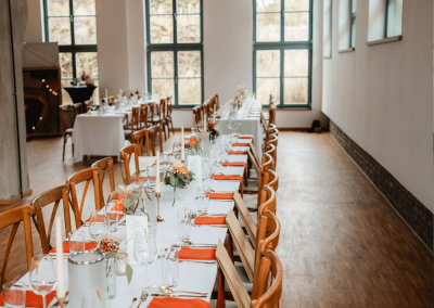 Lange, festlich gedeckte Hochzeitstafel mit Holzstühlen und orangefarbenen Akzenten im Innenraum der Alten Lehrwerkstatt Solingen